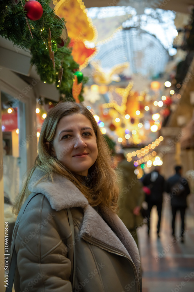 Portrait of serene adult caucasian woman in grey jacket standing in shopping mall with blurred Christmas decorations and garlands in the background. Soft focus. Winter holidays shopping theme. Portrait of serene adult caucasian woman in grey jacket standing in shopping mall with blurred Christmas decorations and garlands in the background. Soft focus. Winter holidays shopping theme.