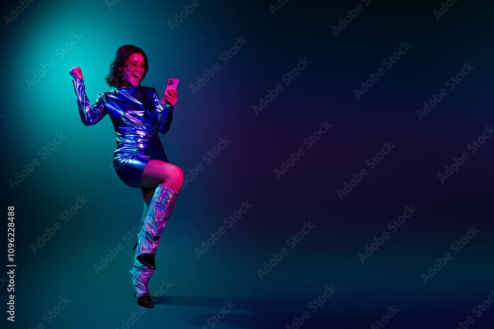Full size photo of gorgeous young girl use phone raise fist empty space wear shiny dress neon light dark blue background Full size photo of gorgeous young girl use phone raise fist empty space wear shiny dress neon light dark blue background