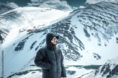 Bearded man in black down jacket and sunglasses standing on a snowy mountain ridge, looking into the distance on a cold winter day