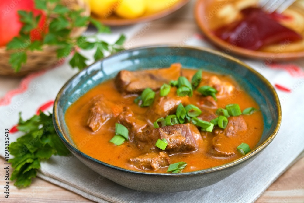 Delicious goulash made from deer meat with green onion in a bowl. Game stew with potatoes and red currant jam at back. Delicious goulash made from deer meat with green onion in a bowl. Game stew with potatoes and red currant jam at back.