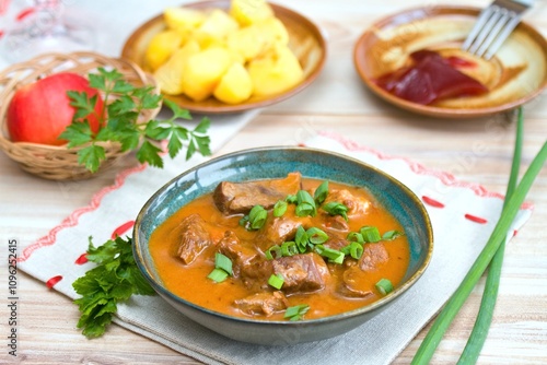 Delicious goulash made from deer meat with green onion in a bowl. Game stew with potatoes and red currant jam at back.