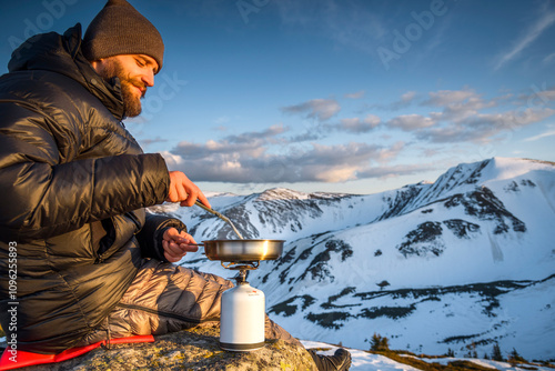 Young hiker with beard cooking on a frying pan during a mountain camping trip at sunset. Concept of escape and solitude