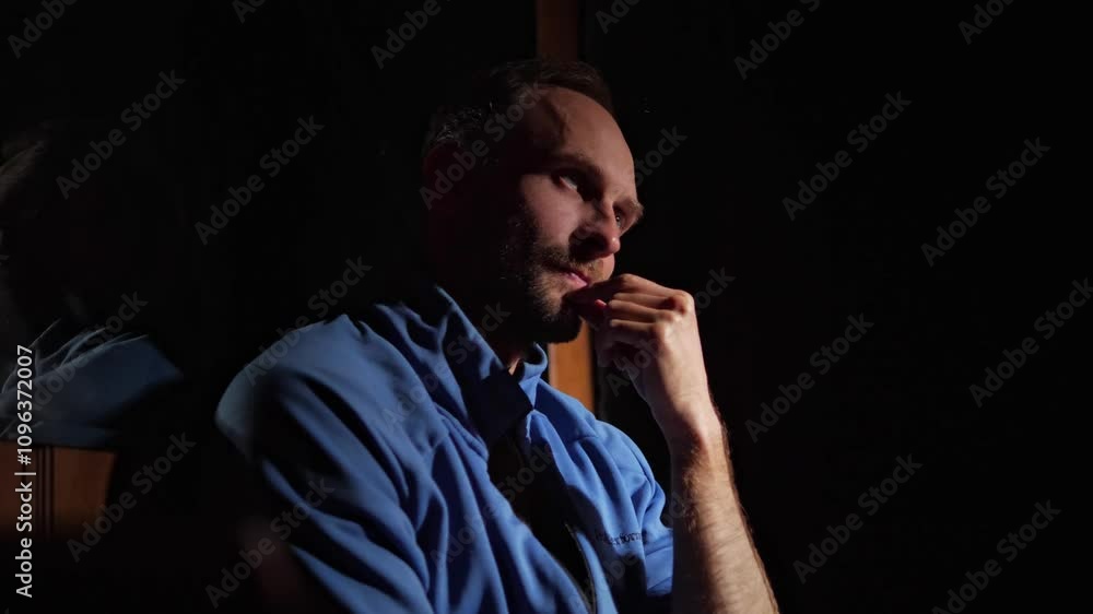 A middle-aged man in a blue shirt sits indoors in a dark, reflective environment, resting his hand on his chin. His serious expression and the contrast between shadows and warm light A middle-aged man in a blue shirt sits indoors in a dark, reflective environment, resting his hand on his chin. His serious expression and the contrast between shadows and warm light