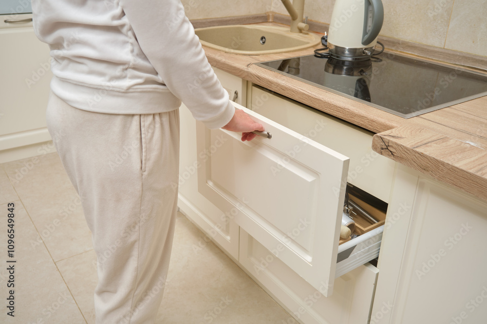 Person opening white kitchen drawer near stove and sink. Close-up view of kitchen interior with beige and wood tones. Home organization and storage concept. Person opening white kitchen drawer near stove and sink. Close-up view of kitchen interior with beige and wood tones. Home organization and storage concept.