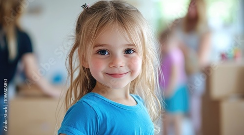 a cute little girl in a blue t-shirt, with blonde hair in a small ponytail, standing and smiling at the camera against a blurred background of a modern living room