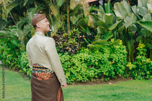 Young Asian man wearing a traditional Javanese beskap and batik standing in a garden. photo