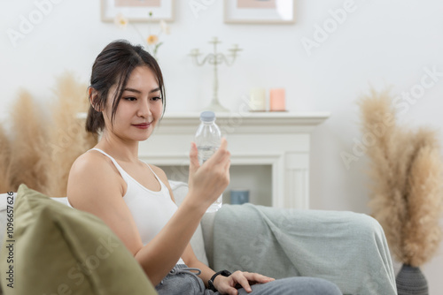 Asian young female sporty holding water bottle sitting on sofa in living room while exercising. stay hydrated per day. Healthy fit woman fresh wellness. Drinking water makes you feel good.