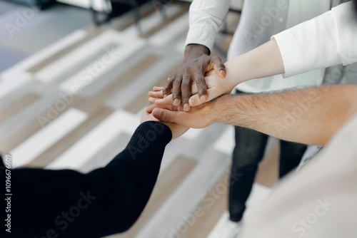 Diverse group joining hands in business environment, symbolizing unity, inclusion, and teamwork among multicultural team members on modern office floor.