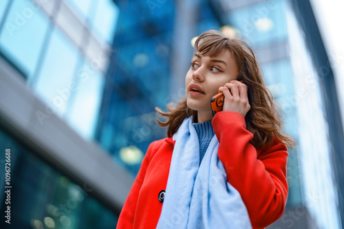 Young woman in red coat talking on phone in modern city setting during daytime