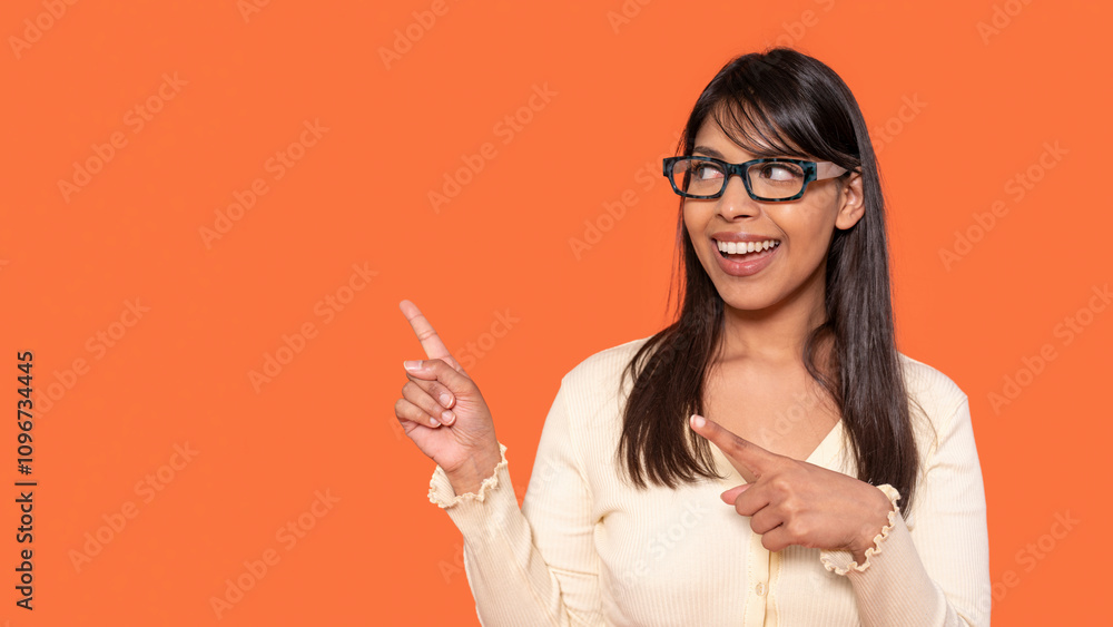 Cheerful woman with glasses pointing at the side against a bright orange background in a casual setting Cheerful woman with glasses pointing at the side against a bright orange background in a casual setting