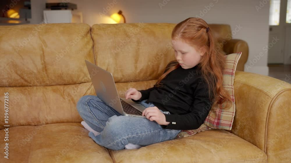 Child using laptop while sitting on couch in cozy living room during afternoon Child using laptop while sitting on couch in cozy living room during afternoon