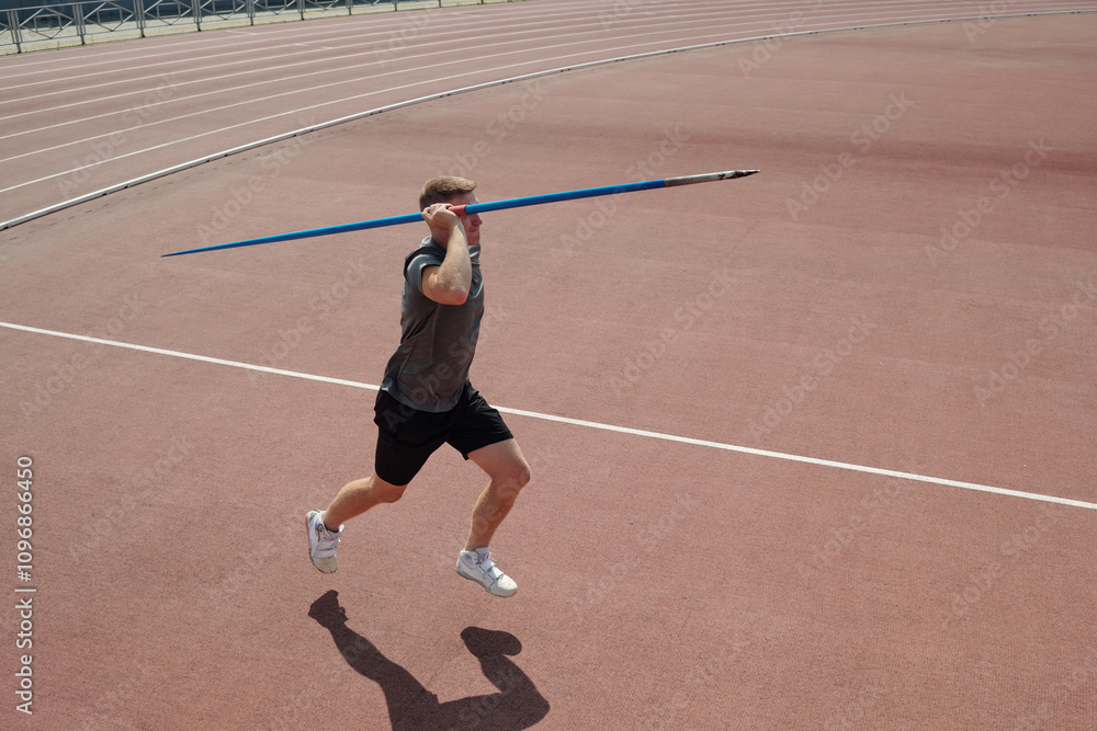 Athlete running on red track field holding javelin above shoulder and preparing release. Background includes part of stadium and fencing surrounding track field Athlete running on red track field holding javelin above shoulder and preparing release. Background includes part of stadium and fencing surrounding track field
