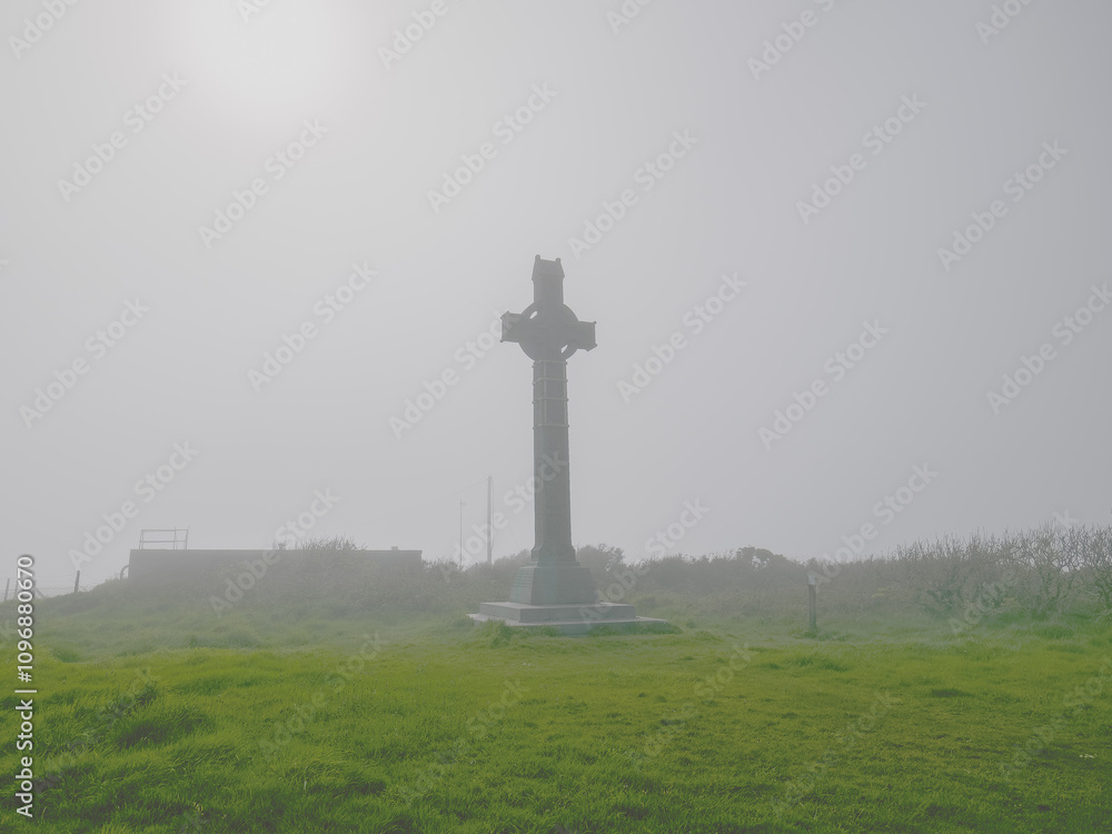A tall Celtic cross is prominently displayed in a lush green field, surrounded by thick fog in the early morning. The atmosphere is tranquil and mystical, creating an ethereal setting. A tall Celtic cross is prominently displayed in a lush green field, surrounded by thick fog in the early morning. The atmosphere is tranquil and mystical, creating an ethereal setting.