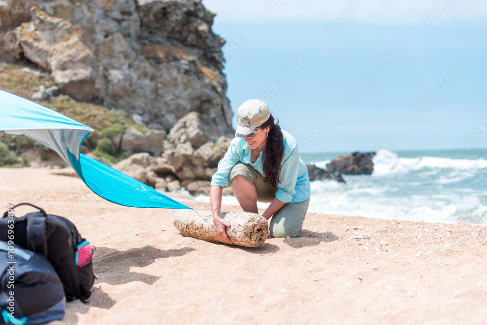 A young slender brunette girl sets up a tent on the seashore to take shelter from the scorching sun. In the background there is a sea and a rock, waves. Tourism. Recreation by savages A young slender brunette girl sets up a tent on the seashore to take shelter from the scorching sun. In the background there is a sea and a rock, waves. Tourism. Recreation by savages
