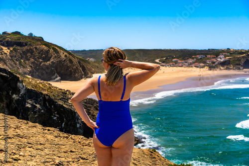 Beautiful mature woman in swimsuit standing on cliff and looking on sunny beach Monte Clerigo, Algarve, Portugal. Back view