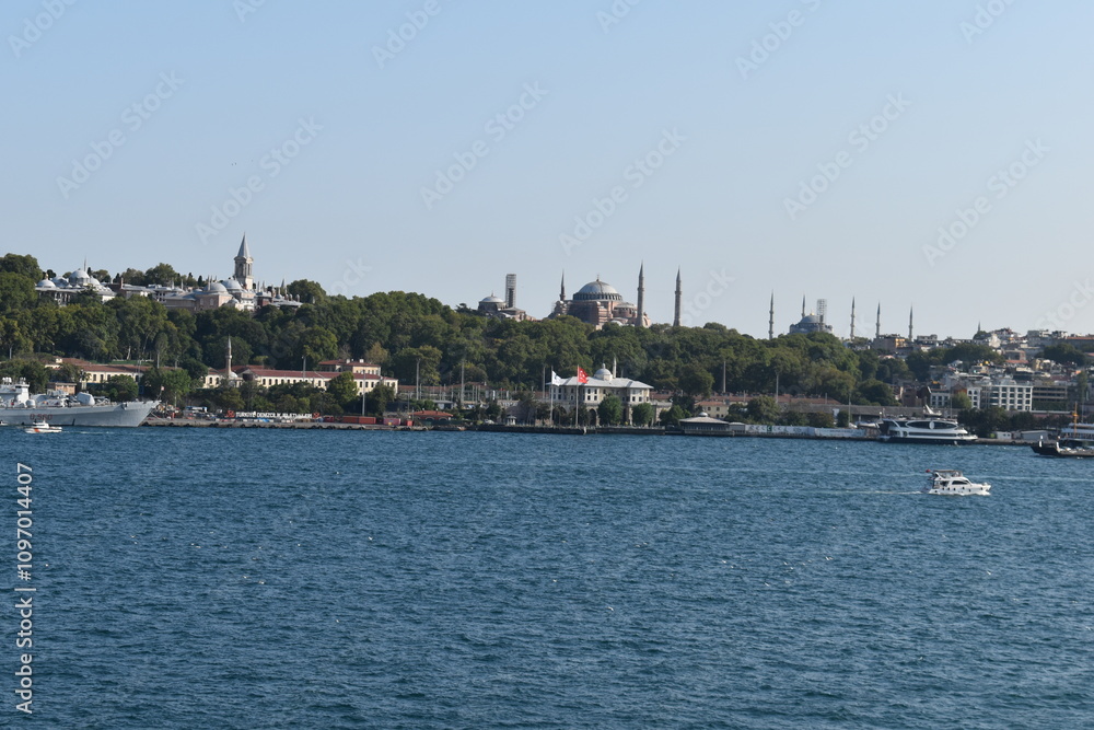 Fototapeta premium A panoramic view of Istanbul's historical peninsula from the Bosphorus Strait, showcasing the Topkapi Palace and Hagia Sophia, surrounded by lush greenery. A small boat navigates the water in the fore