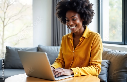 Smiling Black woman uses laptop at home. Young female works remotely. Happy lady chats online. African American person enjoys tech. Modern interior. Freelance job.