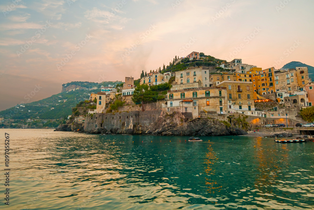 This image showcases Minori on the Amalfi Coast, with its charming seaside houses, tranquil beach, and lush green hills, bathed in the warm Mediterranean light.