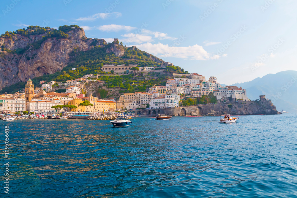 The photo captures a sunny view of Amalfi, showcasing the town's colorful buildings, steep cliffs, and vibrant coastline under a clear blue sky on a bright day.