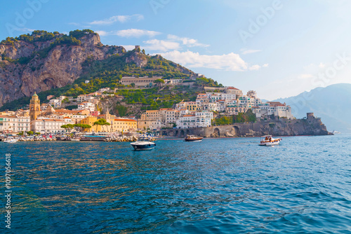 The photo captures a sunny view of Amalfi, showcasing the town's colorful buildings, steep cliffs, and vibrant coastline under a clear blue sky on a bright day.