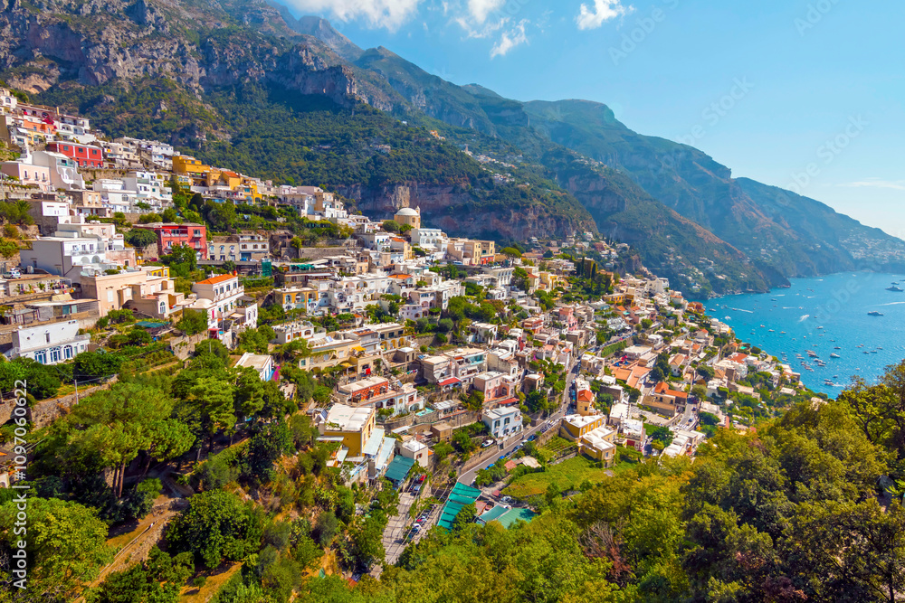 Obraz premium The photo captures a view of Positano, with its colorful, stacked houses cascading down the cliffs, narrow winding streets, and vibrant coastline set against the clear blue sea and sky