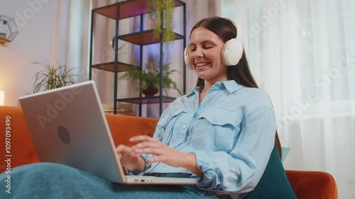 Happy relaxed Caucasian young woman in wireless headphones lying on home couch choosing listening disco music while working online on laptop netbook. Girl weekends activities entertaining. Lifestyles. photo