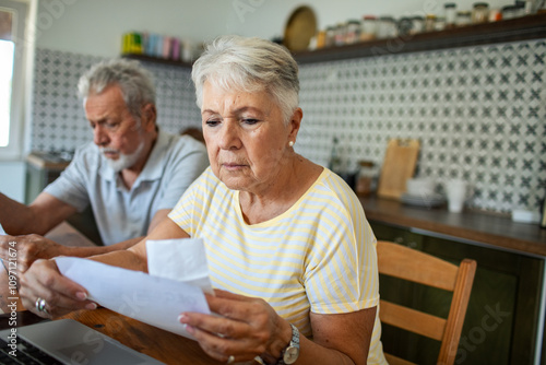 Concerned senior couple reading bills on kitchen table with laptop photo