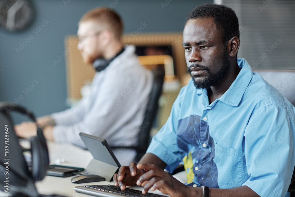 African American man working on laptop in modern office setting filled with light emitting a calm focus, while a colleague is seen in background engaged with his tasks African American man working on laptop in modern office setting filled with light emitting a calm focus, while a colleague is seen in background engaged with his tasks