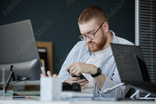 Принти на полотні Man with glasses concentrating while using smartphone and working at office desk
