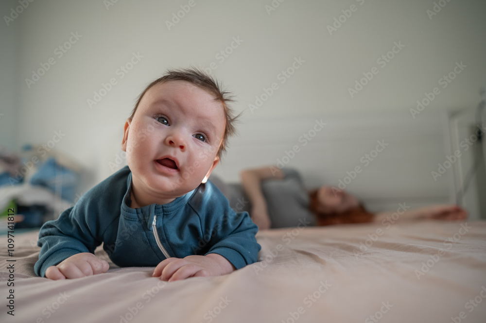 A three-month-old boy lies on his stomach on the bed and his mother sleeps behind him. Postpartum depression. A three-month-old boy lies on his stomach on the bed and his mother sleeps behind him. Postpartum depression.