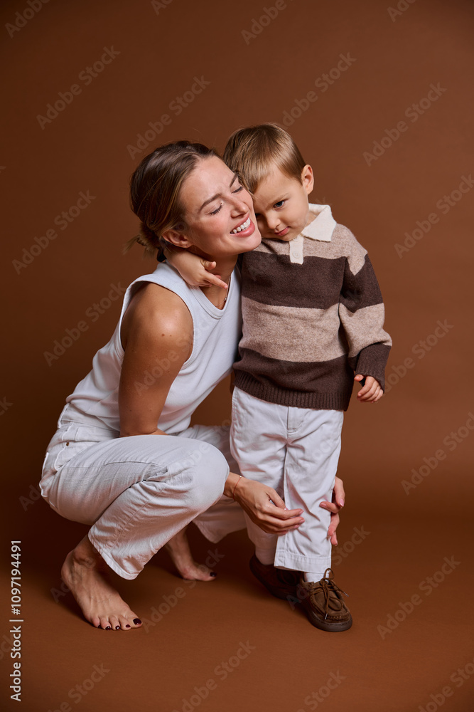 A mother and her young son enjoy a joyful moment together in a studio. The mother kneels close, smiling affectionately at her son, who is playfully leaning against her. A mother and her young son enjoy a joyful moment together in a studio. The mother kneels close, smiling affectionately at her son, who is playfully leaning against her.