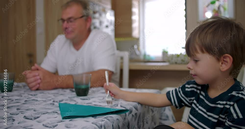 Boy sitting at table with family nearby, engaged in quiet and natural interaction, authentic glimpse into daily family routine in a warm kitchen setting Boy sitting at table with family nearby, engaged in quiet and natural interaction, authentic glimpse into daily family routine in a warm kitchen setting