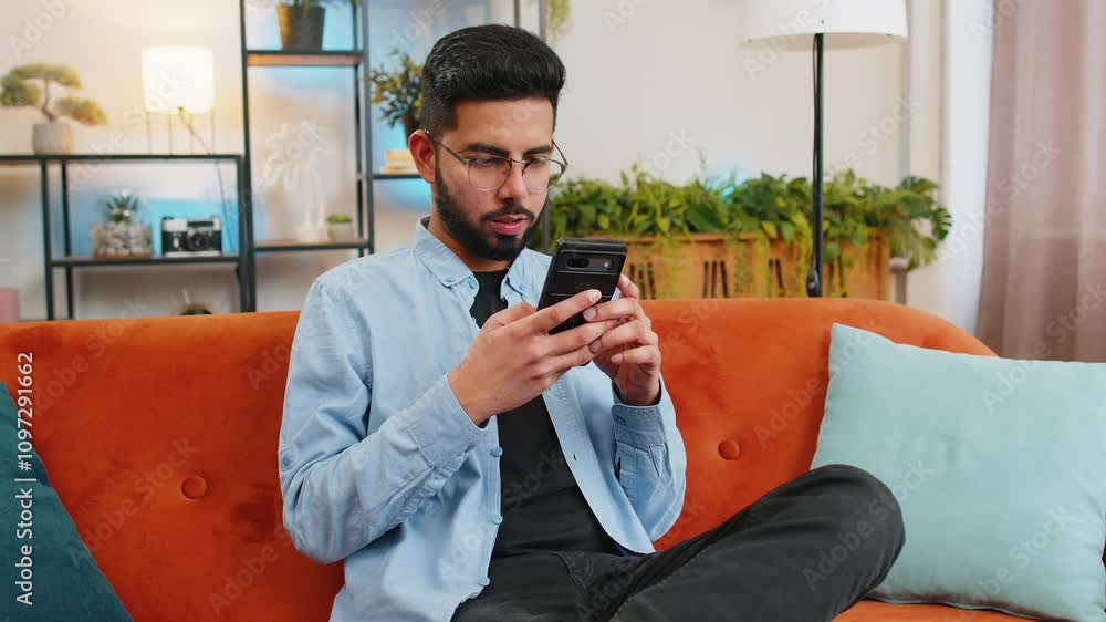 Happy young man in casual clothes thinking and text messaging on smartphone. Indian guy sitting on sofa and using cellphone in living room at home. Male is searching information on browser internet.