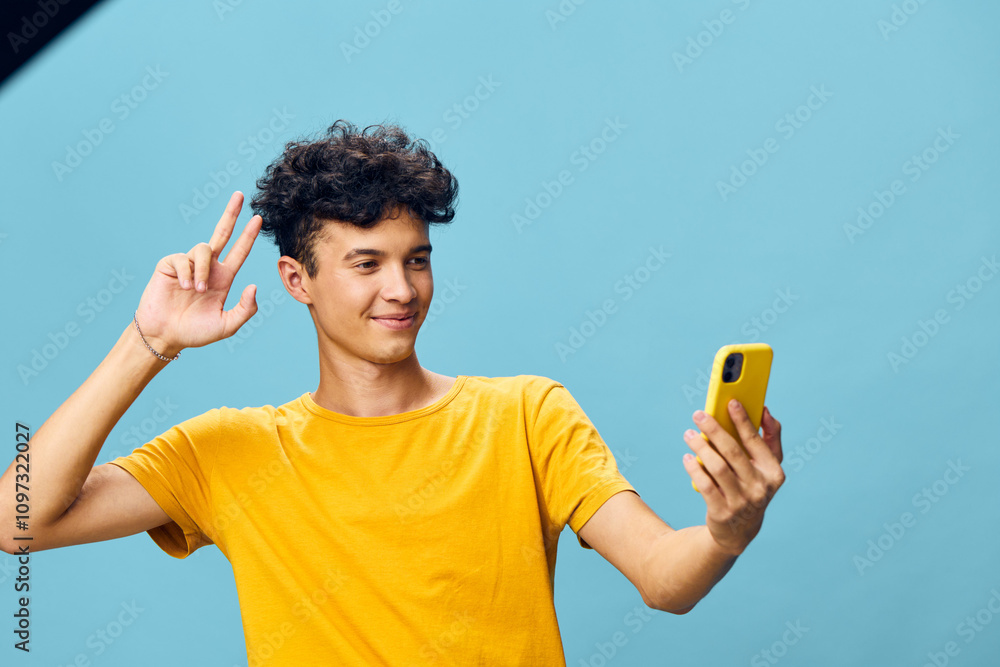 Young man taking a selfie with a smartphone against a light blue background, smiling and making a peace sign, showcasing modern technology and social interaction Young man taking a selfie with a smartphone against a light blue background, smiling and making a peace sign, showcasing modern technology and social interaction