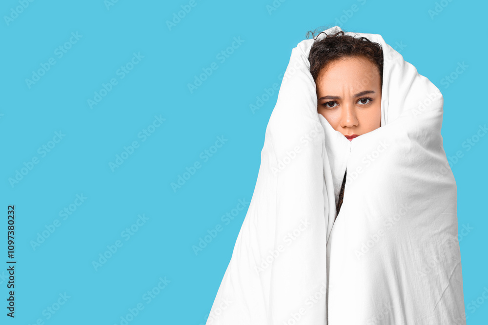 Beautiful young displeased African-American woman in pajamas with blanket on blue background Beautiful young displeased African-American woman in pajamas with blanket on blue background