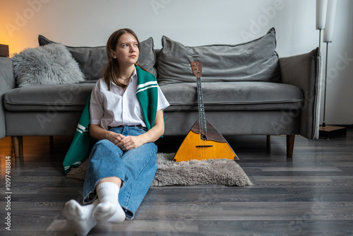 Thoughtful young woman resting after playing on Russian balalaika at home, sitting on faux fur rug on the floor, thinking and looking at window. Folk stringed musical instrument. Hobby. 