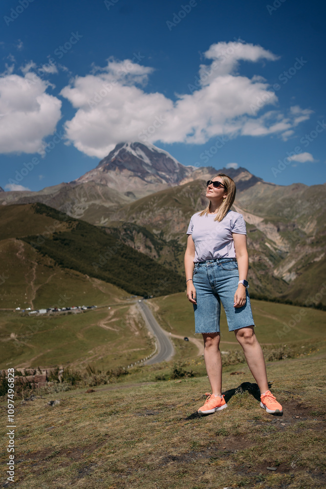 Fototapeta premium A tourist girl on the background of Mount Kazbek in Georgia near the village of Stepantsminda. The girl is wearing a purple T-shirt, blue denim shorts, sunglasses. There is a mountain with a snow cap 