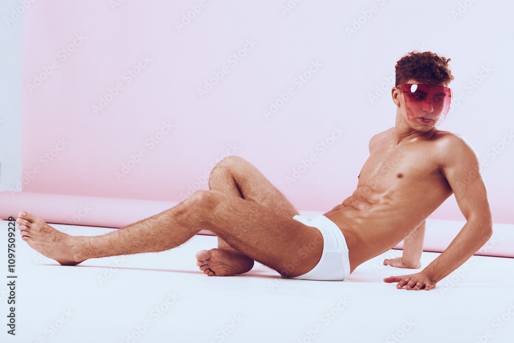 Obraz premium Stylish young man lounging in minimalistic setting, wearing sunglasses and sporty white shorts, showcasing confidence and modern fashion.