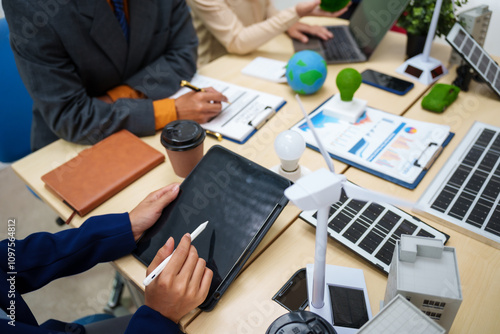 Asian business professionals in collaborate at desk with building models, planning sustainable projects solar panels, electric cars, wind turbines, eco-friendly practices and clean energy solutions.