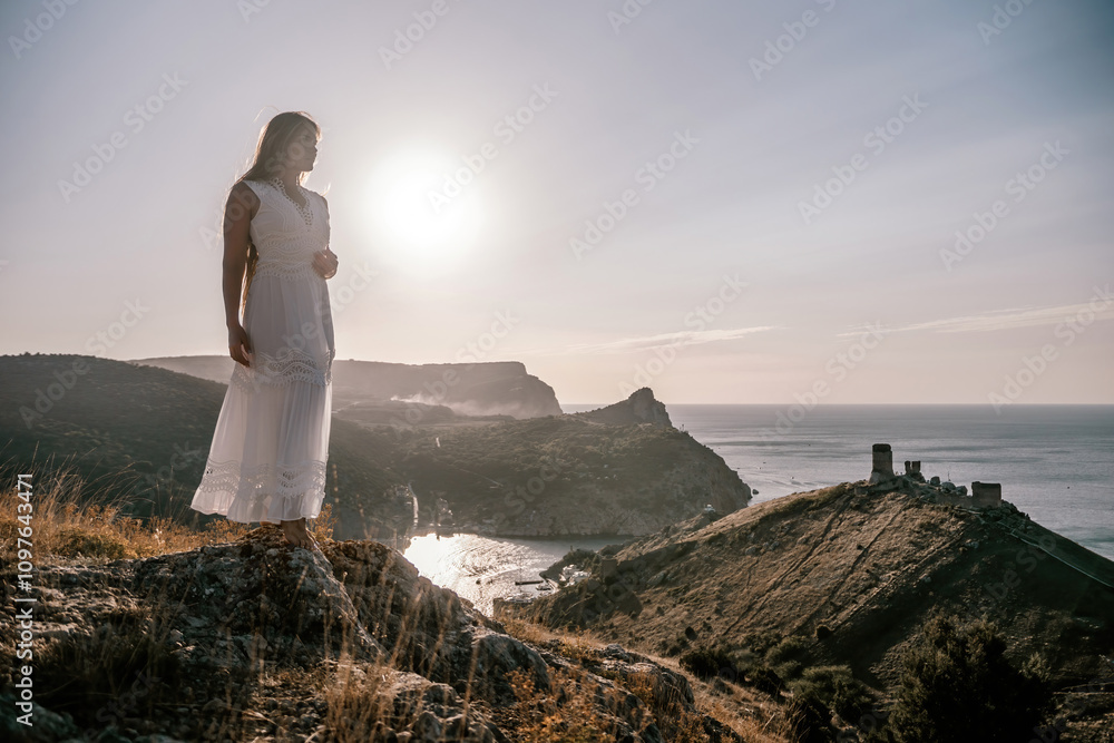 A woman stands on a rocky hill overlooking the ocean. She is wearing a white dress and she is enjoying the view. The scene is serene and peaceful, with the sun shining brightly in the background. A woman stands on a rocky hill overlooking the ocean. She is wearing a white dress and she is enjoying the view. The scene is serene and peaceful, with the sun shining brightly in the background.