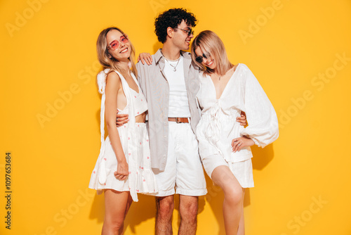 Group of young three stylish friends posing in studio. Fashion man and two cute female dressed in casual summer clothes. Smiling models having fun. Cheerful women and guy near yellow wall, sunglasses