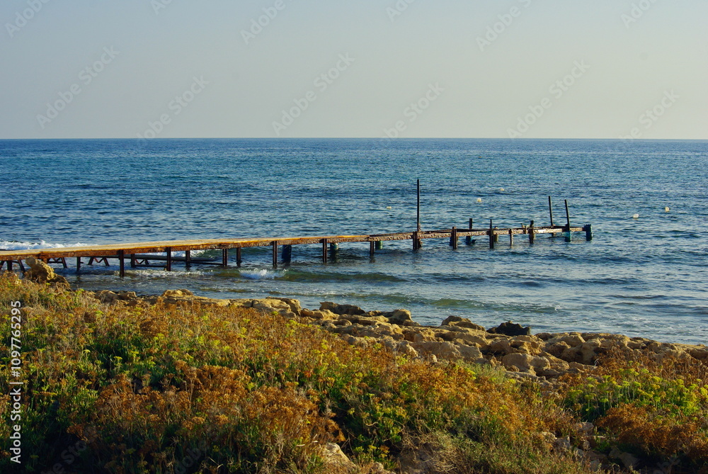 A scenic wooden pier extending into the blue Mediterranean Sea in Ayia Napa, Cyprus, surrounded by rocky shores and coastal vegetation A scenic wooden pier extending into the blue Mediterranean Sea in Ayia Napa, Cyprus, surrounded by rocky shores and coastal vegetation