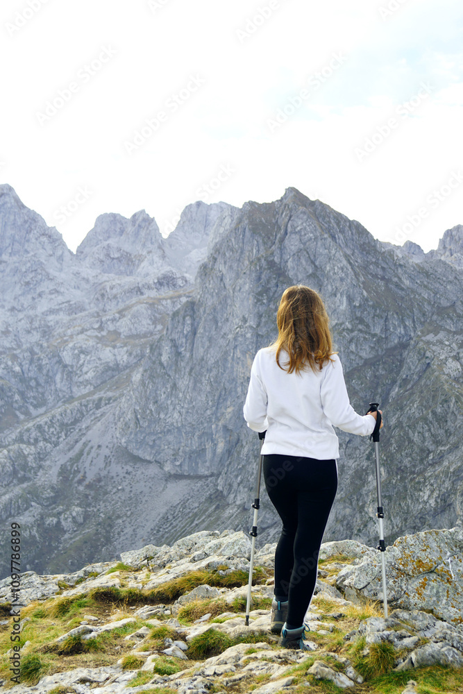 Naklejka premium A girl in sportswear stands leaning on hiking boards at the top and enjoys the view of the mountains. Walking the trail between Volusnica and Talijanka peaks in Prokletije National Park, Montenegro.
