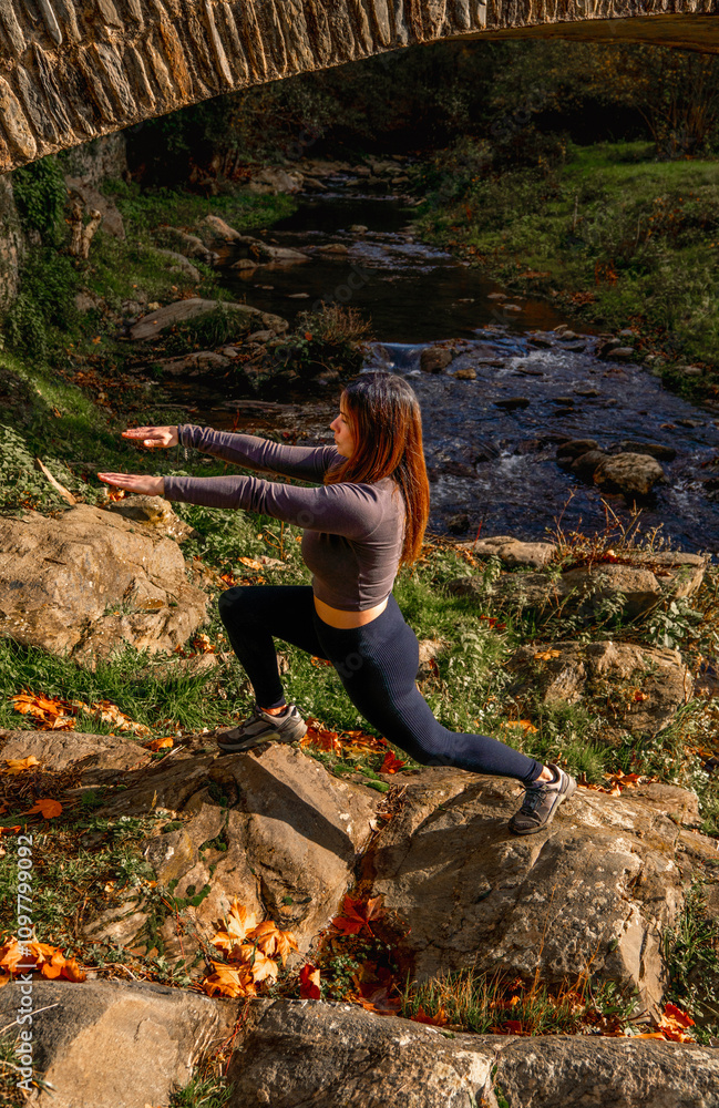 Obraz premium Young woman in tight sportswear performing a lunge exercise on rocks next to a mountain river near a stone bridge in an autumn landscape.