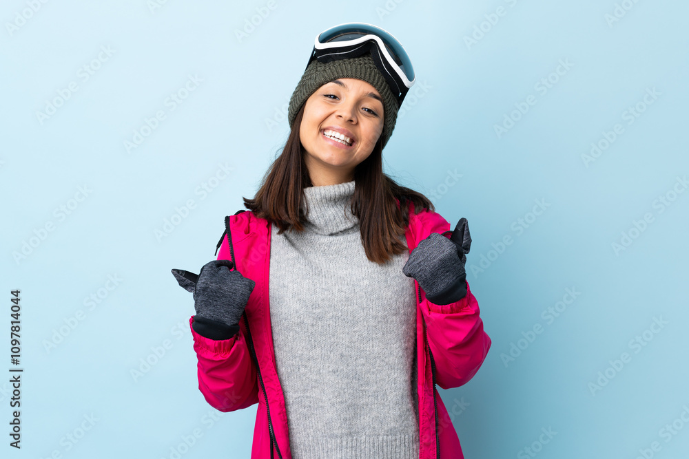 Mixed race skier girl with snowboarding glasses over isolated blue background with thumbs up gesture and smiling. Mixed race skier girl with snowboarding glasses over isolated blue background with thumbs up gesture and smiling.