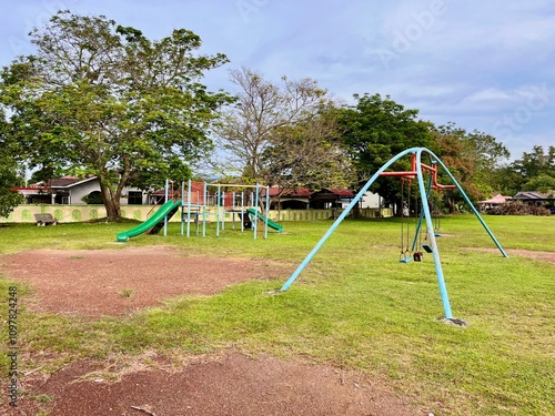 A vibrant playground with swings, slides, and seesaws in a lush residential park, surrounded by shady trees and clean walking paths.