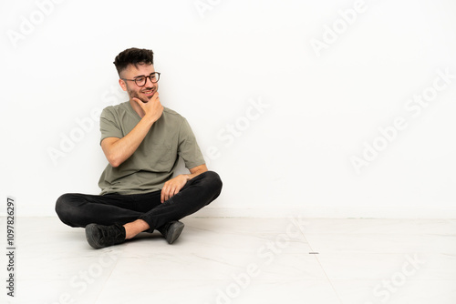 Young caucasian man sitting on the floor isolated on white background looking to the side