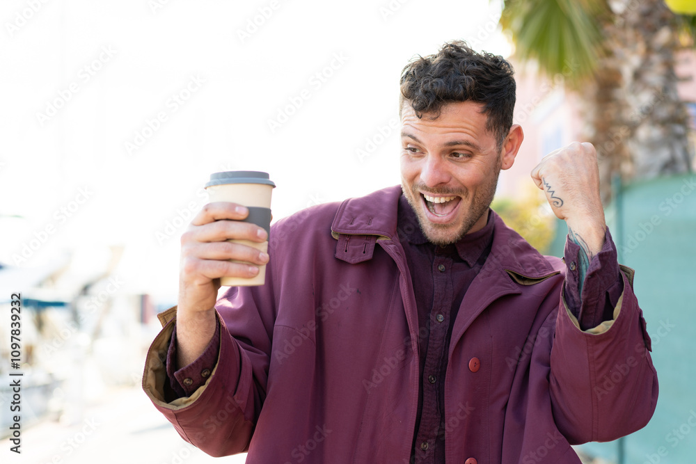 Young caucasian man holding a take away coffee at outdoors celebrating a victory Young caucasian man holding a take away coffee at outdoors celebrating a victory