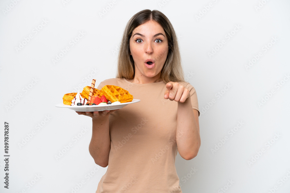 Young Rumanian woman holding waffles isolated on white background surprised and pointing front Young Rumanian woman holding waffles isolated on white background surprised and pointing front