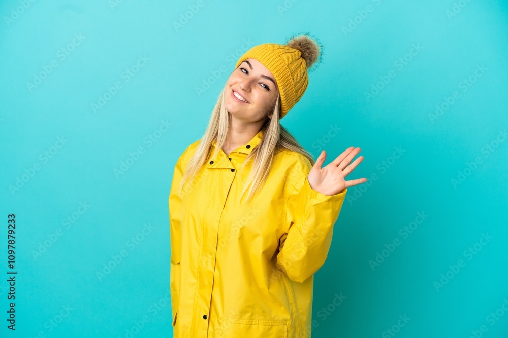 Young woman wearing a rainproof coat over isolated blue background saluting with hand with happy expression Young woman wearing a rainproof coat over isolated blue background saluting with hand with happy expression